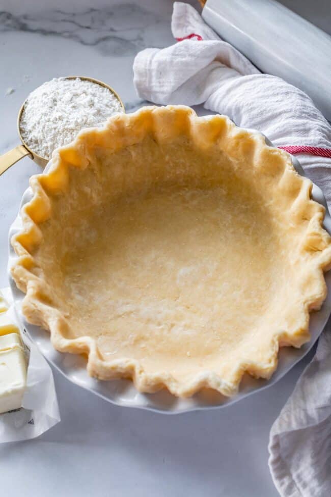 Fluted single easy flaky pie crust in a pan with flour, sliced unwrapped stick of butter, and rolling pin.