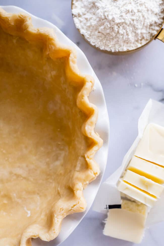 buttery flaky pie crust in a white ceramic pie pan next to a sliced, partially unwrapped stick of butter shot from above.
