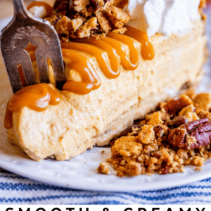 A slice of no-bake pumpkin cheesecake topped with whipped cream, caramel sauce, and pecan crumble, with a fork cutting in. A pumpkin and blurred background complete the cozy, autumn-inspired scene.