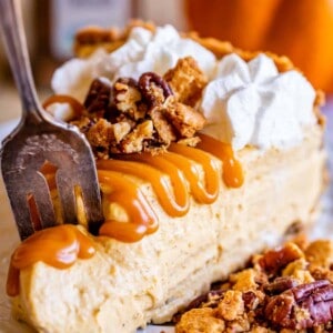 A slice of creamy no bake pumpkin cheesecake topped with whipped cream, caramel sauce, and pecan crumble sits on a plate as a fork cuts in; a blurred pumpkin is in the background.