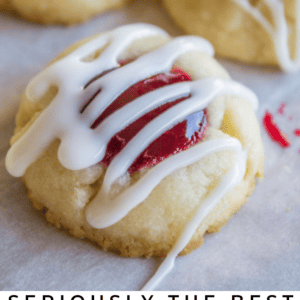Close-up of raspberry thumbprint cookies filled with raspberry jam and topped with a white icing drizzle, set on parchment paper. Text below reads “Seriously Delicious Raspberry Thumbprint Cookies.”.