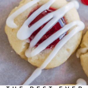Close-up of raspberry thumbprint cookies drizzled with white icing, each cookie filled with vibrant red raspberry jam, on a light surface. The text below reads: Raspberry Thumbprint Cookies.