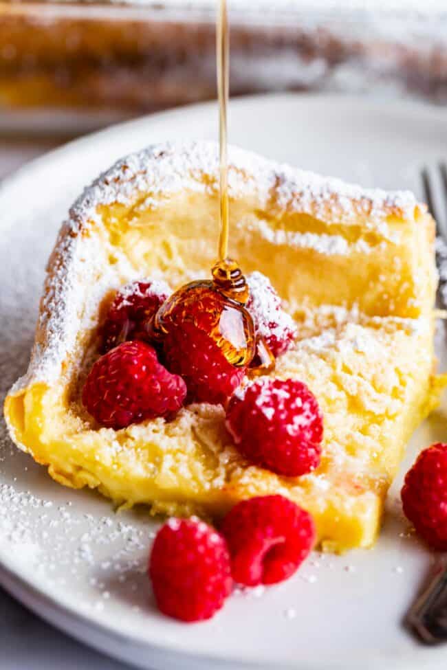 dutch baby pancake on a plate with raspberries and powdered sugar, being drizzled with syrup.