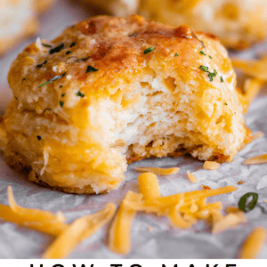 A close-up of a golden, cheesy biscuit with a bite taken out, topped with herbs, surrounded by shredded cheddar and similar biscuits in the background. Text reads: “Cheddar Bay Biscuits.”.