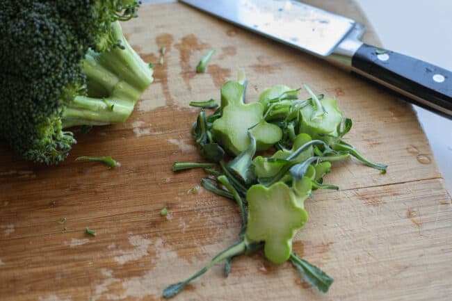 peeling leaves off of broccoli stems, chopping of end of broccoli stems on a wooden cutting board