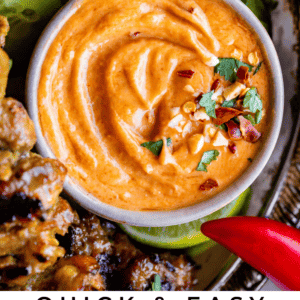 A bowl of creamy Thai peanut sauce garnished with herbs and crushed peanuts sits on a platter with sliced cucumbers and grilled meat. Text reads: Quick & Easy Thai Peanut Sauce - The Food Charlatan.