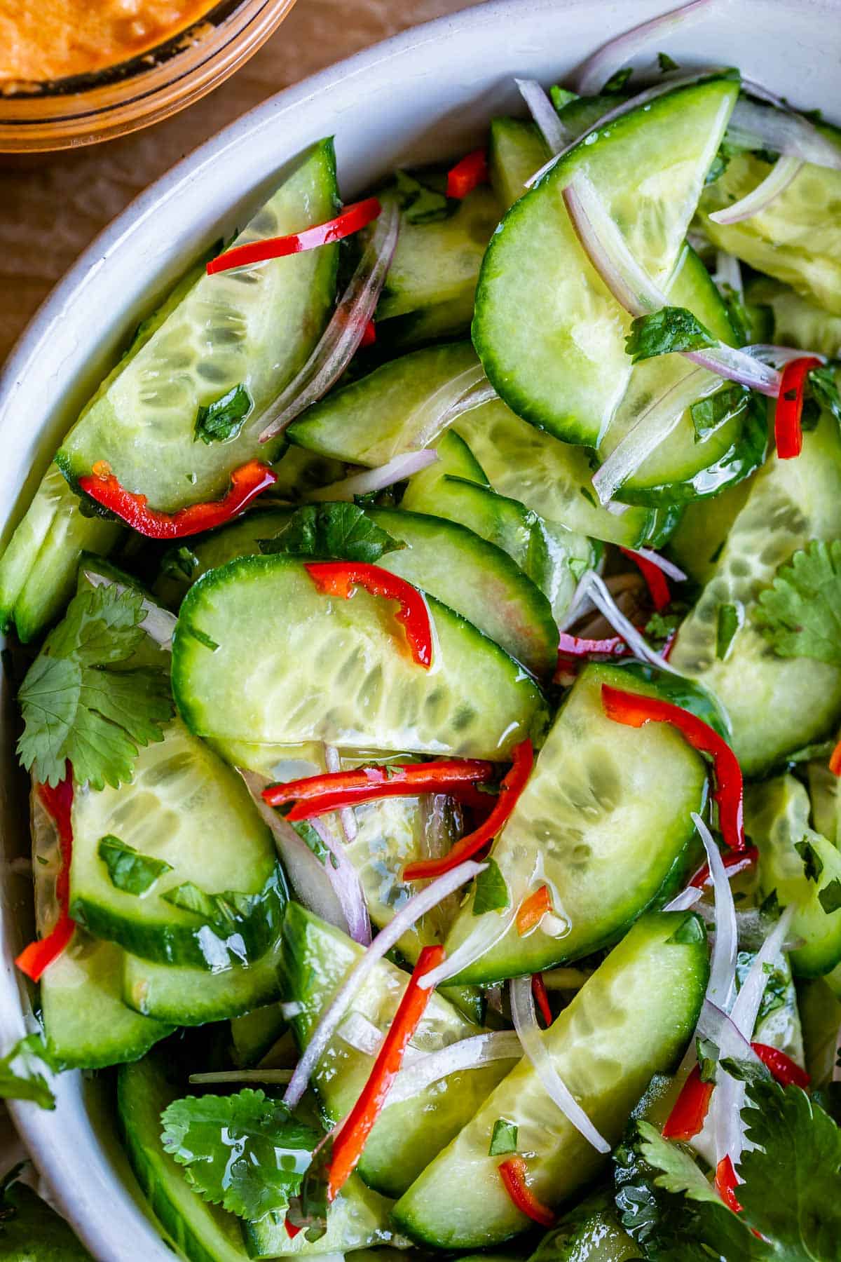 Thai Cucumber Relish for Satay 12 Close-up of a vibrant Thai Cucumber Salad in a white bowl, featuring thinly sliced red peppers and shallots among crisp cucumbers, bathed in a clear vinegar syrup.