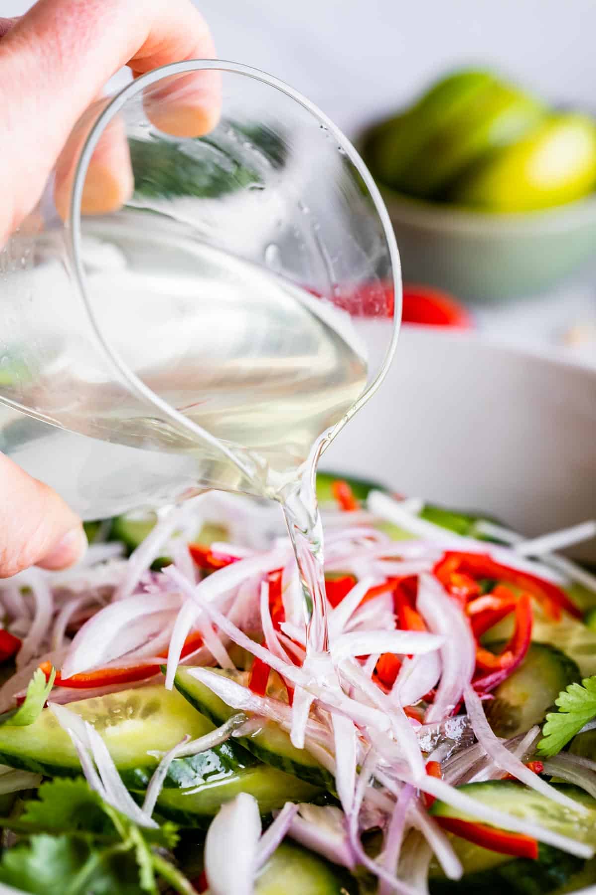 Thai Cucumber Relish for Satay 9 A stream of clear vinegar syrup being poured over a colorful mix of thinly sliced shallots, red chili peppers, and crisp cucumbers in a white ceramic bowl, illustrating the final step of preparing Thai Cucumber Salad.