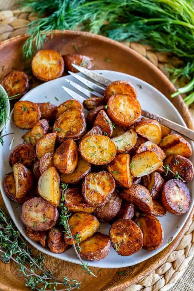 oven roasted red potatoes on a white plate placed on a wooden plate, with dill on the side.