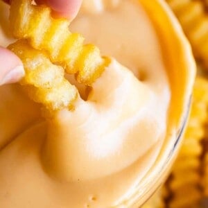 two crinkle fries being dipping into a clear bowl of easy cheese sauce.