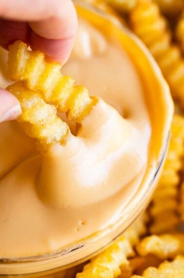two crinkle fries being dipping into a clear bowl of easy cheese sauce.