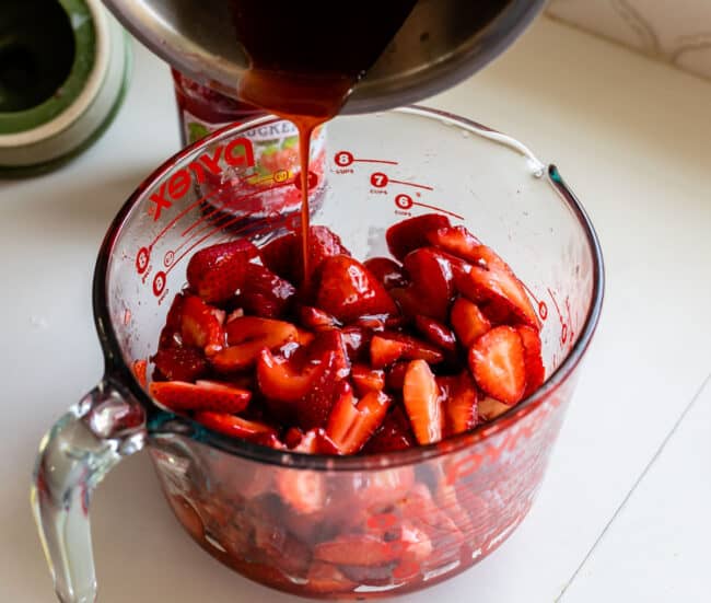 adding cooked jam to a bowl of sliced strawberries.