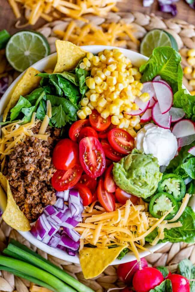 Bowl of taco salad with limes, corn, guacamole, meat, cheese, chips, tomatoes, and jalapenos. 