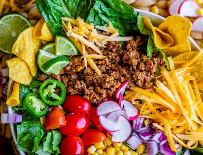 a bowl of taco salad ingredients like lettuce, meat, cheese, chips, tomatoes, limes, and more.