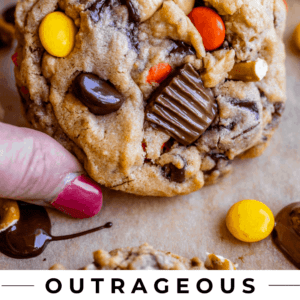 A hand holds a thick, chewy cookie packed with pretzels, chocolate chunks, and Reeses Pieces. Other cookies and candy pieces are visible in the background. Text reads: Outrageous Reeses Peanut Butter Cookies. The Food Charlatan.