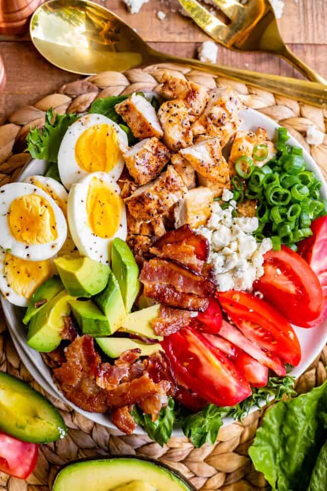 a Cobb salad with chicken, bacon, cheese, eggs, and vegetables next to gold salad utensils.