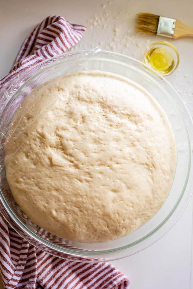 pizza dough rising in a glass bowl with plastic wrap.