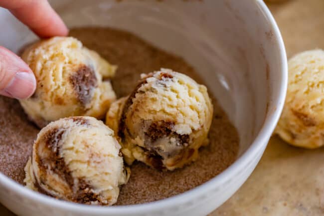 cinnamon roll cookie dough being rolled in cinnamon sugar in a white bowl