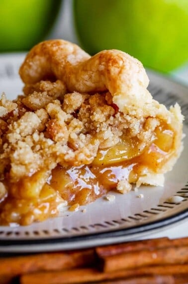 A close-up of a Dutch apple pie slice with crumb topping on a plate, showing gooey apple filling, golden crust, and crumbly topping; green apples and cinnamon sticks are in the background.