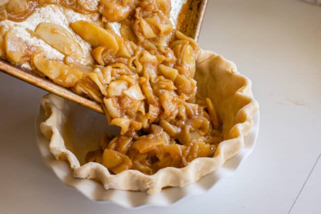 pouring chilled apple pie filling from a sheet pan into a frozen pie crust