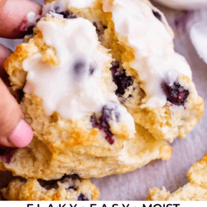 A hand holds a flaky scone with white icing and visible berries, surrounded by more scones and a bowl of red jam. Text at the bottom reads Flaky, Easy, Moist Berry Scones, The Food Charlatan.