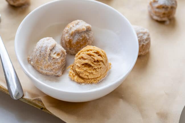 peanut butter cookie dough balls being rolled in a bowl of sugar.