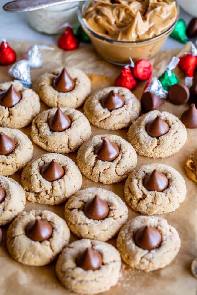 peanut butter blossoms on parchment paper with Hershey's kisses and a bowl of peanut butter in the background.
