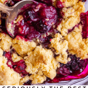 A close-up of a baked berry cobbler with a golden, crumbly topping and vibrant fruit filling, with a large spoon scooping some out. Text reads “Seriously Delicious Berry Cobbler - The Food Charlatan.”.