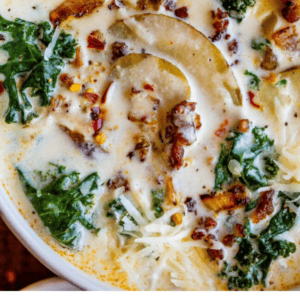 A close-up of creamy Zuppa Toscana soup with sausage, kale, potato slices, cheese, and spices, served in a white bowl with a spoon. Text overlay reads Zuppa Toscana sausage & kale soup. The Food Charlatan.