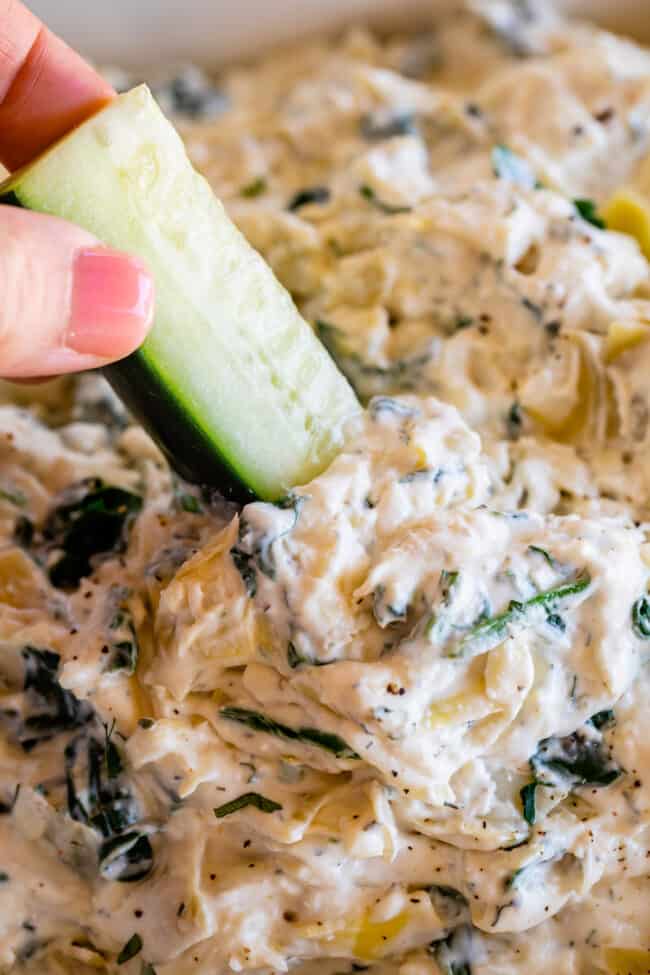 hand dipping cucumber spear into spinach and artichoke dip