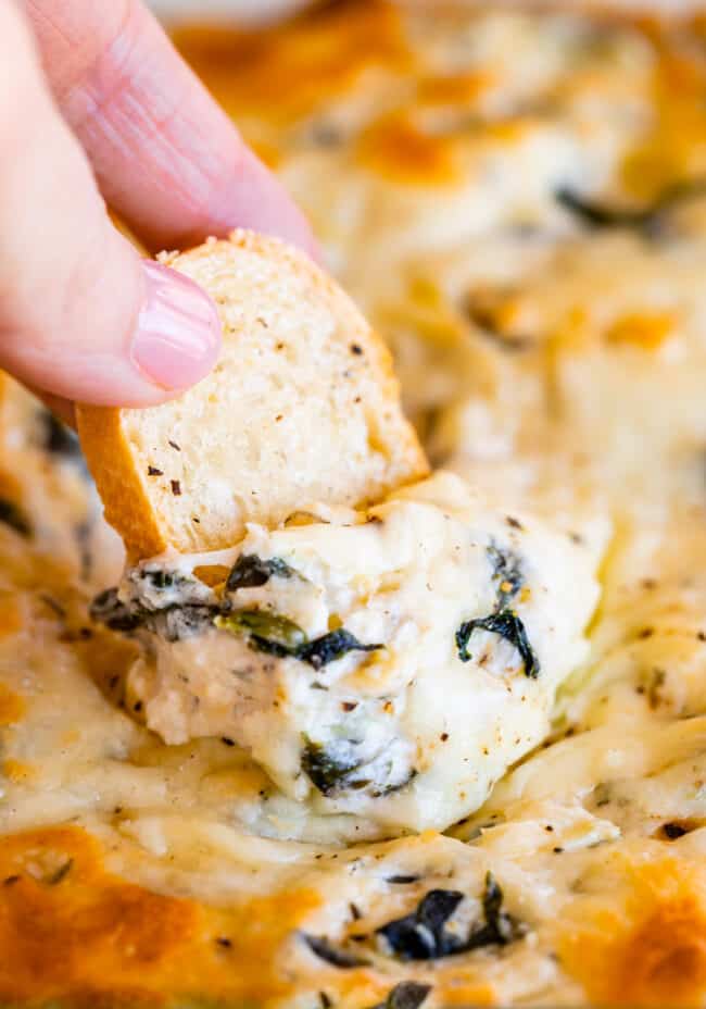 hand dipping toasted baguette into hot spinach artichoke dip