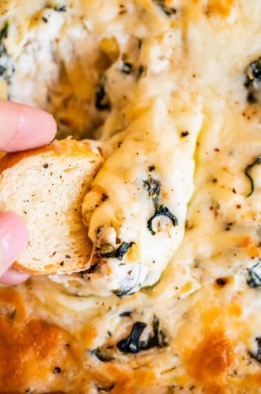 hand dipping bread into cheesy best spinach artichoke dip