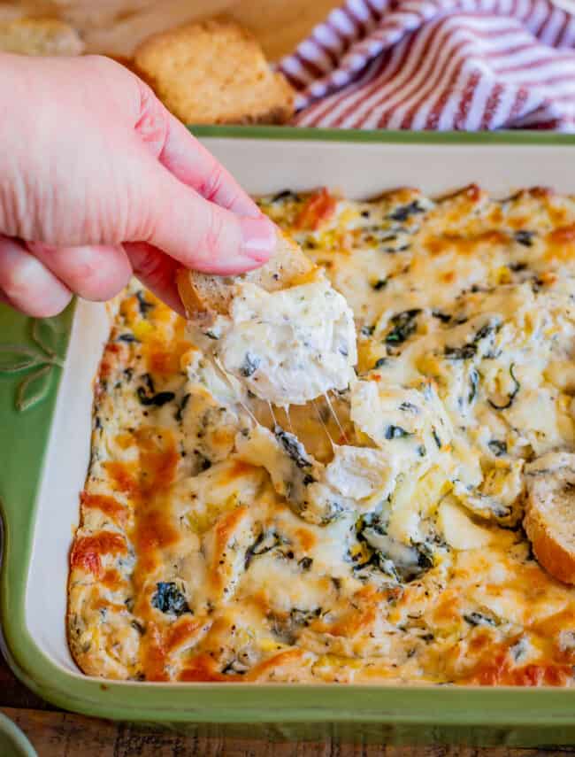 hand dipping toasted bread into a pan of best spinach artichoke dip