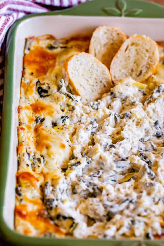 spinach and artichoke dip in a green casserole dish with 3 toasted bread slices