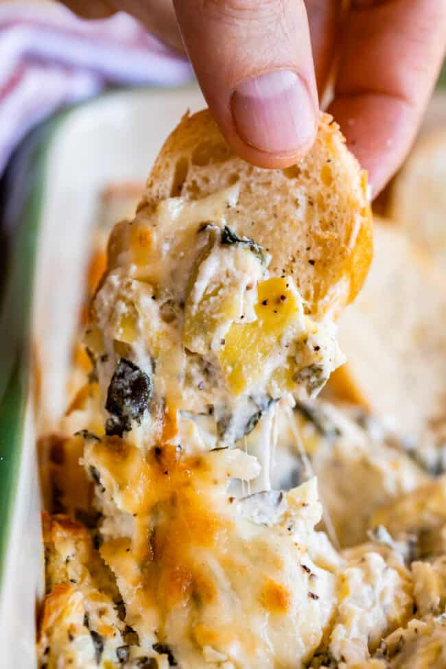 hand dipping bread into hot spinach artichoke dip with cheese pull