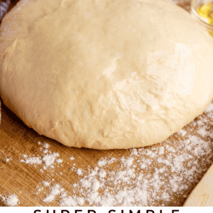 A ball of pizza dough rests on a floured wooden surface with a red and white striped cloth and a small bowl of olive oil nearby. Text reads: Super Simple Beginners Pizza Dough with olive oil - The Food Charlatan.