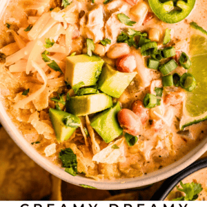 A bowl of creamy white chicken chili topped with shredded cheese, avocado cubes, jalapeño slices, green onions, and cilantro, served with a lime wedge. Text below reads: Creamy Dreamy White Chicken Chili - The Food Charlatan.