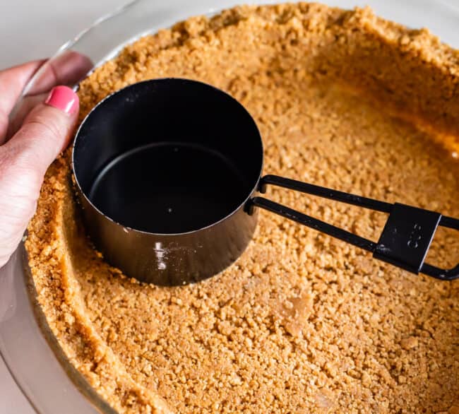 pressing graham cracker crumbs into a pie pan using a black measuring cup.