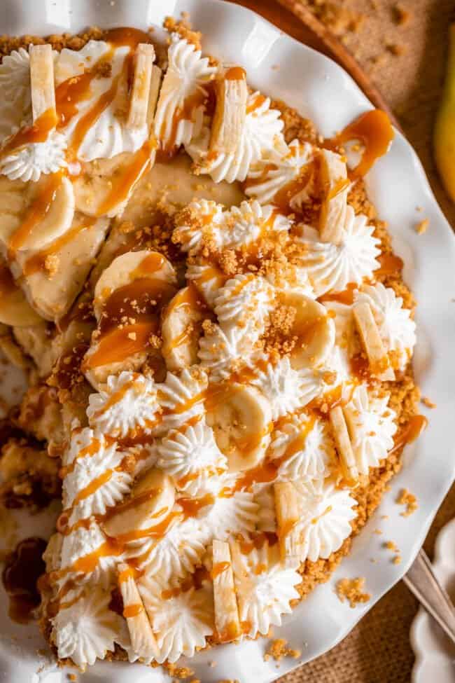 banana cream pie in a white pie plate drizzled with caramel and graham crumble.