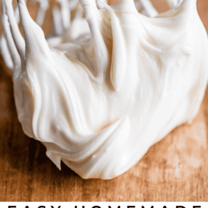 A metal whisk covered in creamy, thick white frosting rests on a wooden surface. Text at the bottom reads: Easy Cream Cheese Frosting. The Food Charlatan.