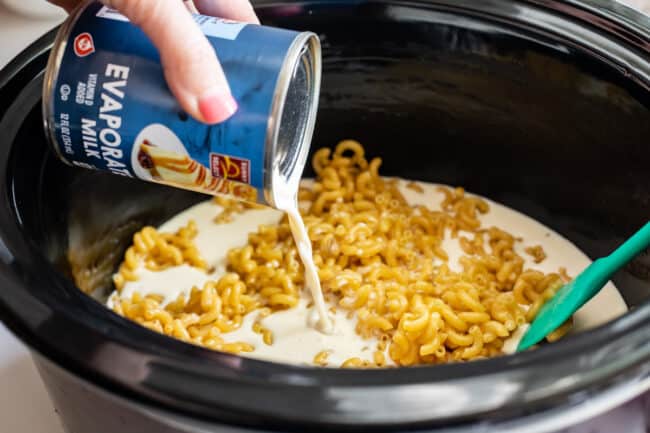 adding a can of evaporated milk to a crock pot full of dry noodles.