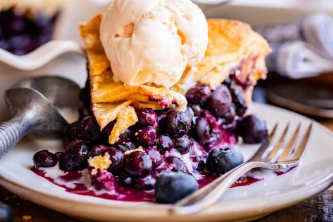 blueberry pie recipe on a plate with fork and ice cream scoop, topped with ice cream.