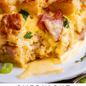 A close-up of a slice of eggs benedict casserole with chunks of bread, ham, cheese, and fresh herbs, topped with creamy hollandaise sauce on a white plate. Text reads: Overnight Eggs Benedict Casserole, Brunch Inspiration, The Food Charlatan.