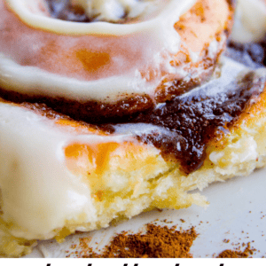 Close-up of a cinnamon roll topped with white icing on a plate, with text overlay at the bottom that reads “truly irresistible cinnamon rolls - The Food Charlatan.”.