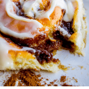 A close-up of a frosted cinnamon roll on a plate with a fork, partially eaten, revealing gooey cinnamon filling. Text below reads: Irresistibly gooey cinnamon rolls - The Food Charlatan.