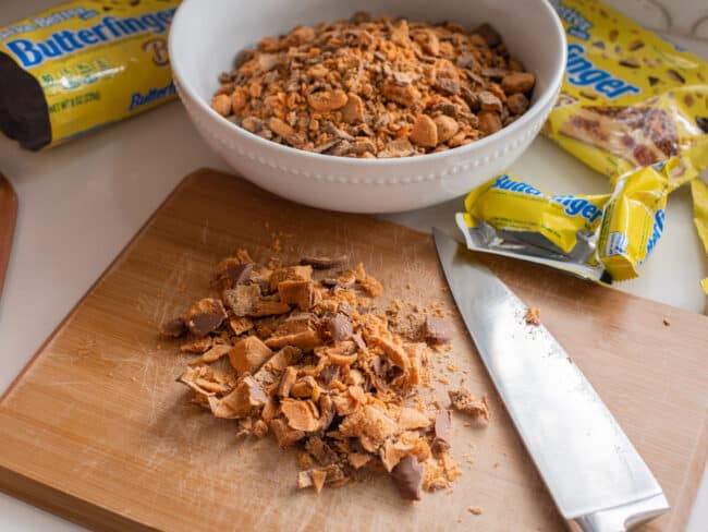chopped up butterfingers on a cutting board with a bowl of butterfinger bits in the background.