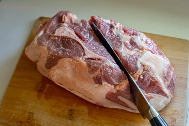 A chef's knife splitting a pork should in half on a cutting board.