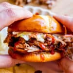 hands holding smoked bbq pulled pork on a bun with white plate and chips.