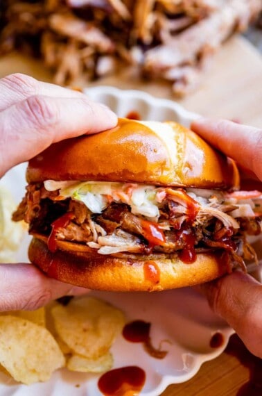 hands holding smoked bbq pulled pork on a bun with white plate and chips.