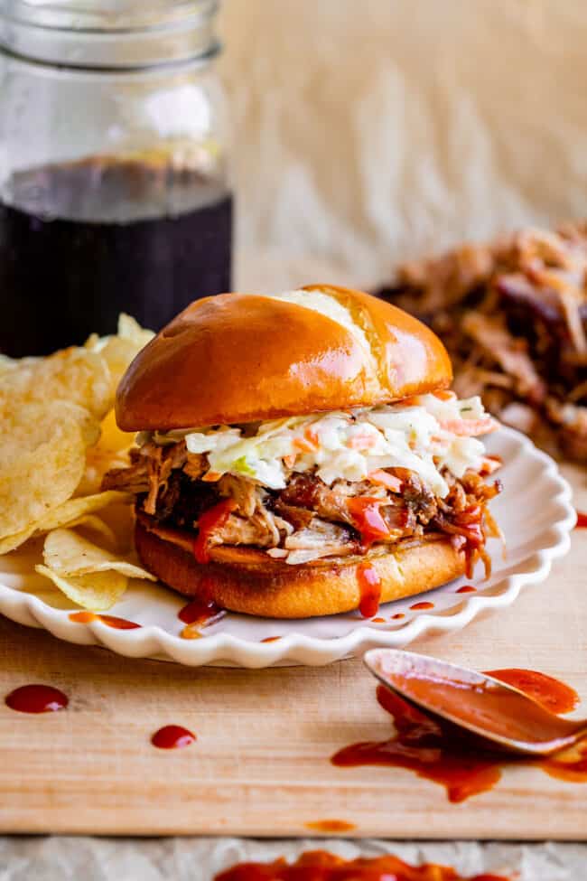 smoked pulled pork sandwich with coleslaw on a white plate with soda and chips.
